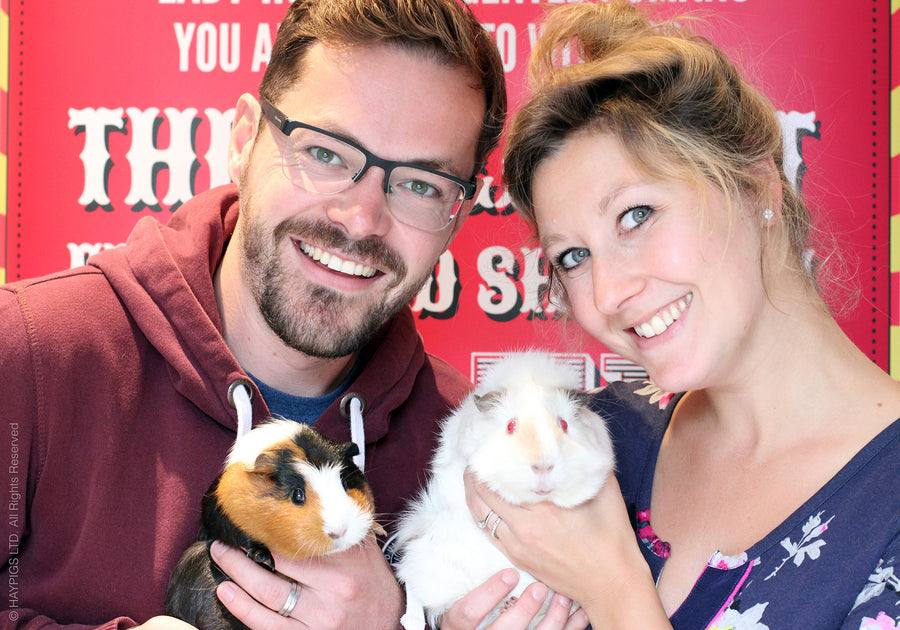 Rik and Helen Cridland, co-founders of HayPigs!, each holding a guinea pig in front of a red circus-style banner